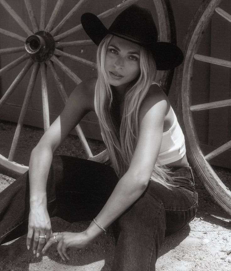 Woman in a cowboy hat and tank top sitting outdoors next to wagon wheels