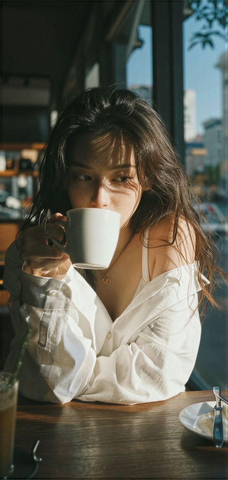 Woman in a white shirt drinking from a mug with dappled sunlight on her face