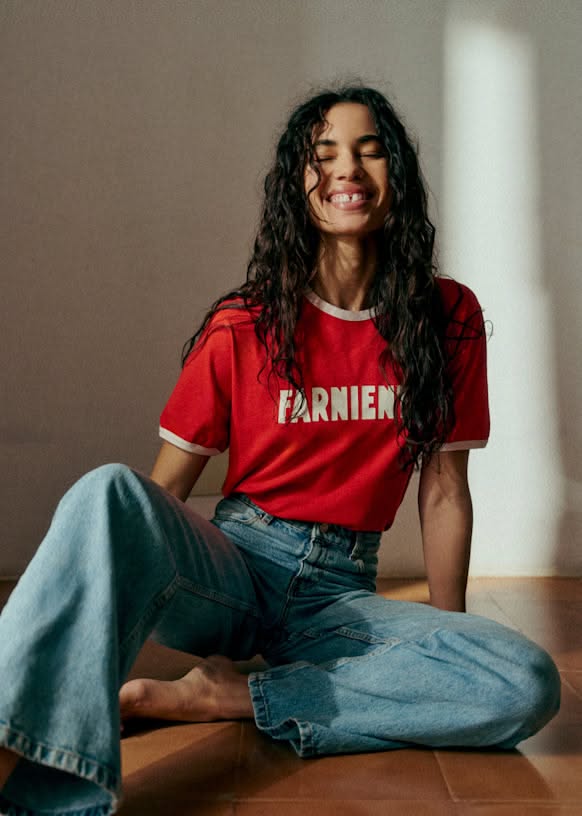 Woman with long curly hair in a red t-shirt and blue jeans sitting on a wooden floor