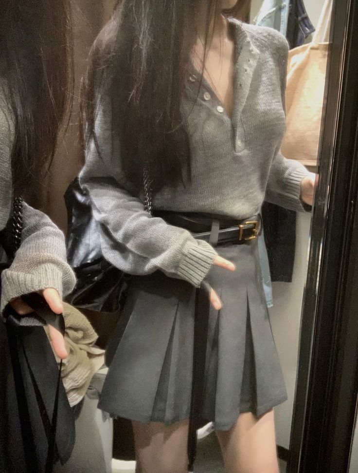 Person in a grey sweater and pleated skirt reflected in a mirror