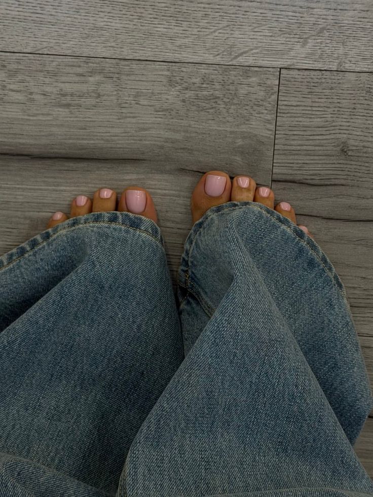 Bare feet with pink nail polish peeking out from light wash jeans on a wooden floor