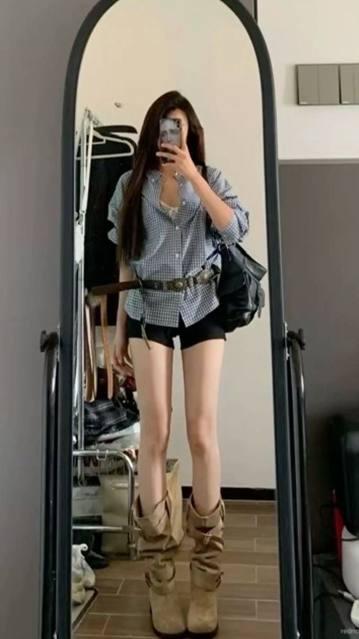 Woman in a gingham shirt, black shorts, and cowboy boots taking a mirror selfie