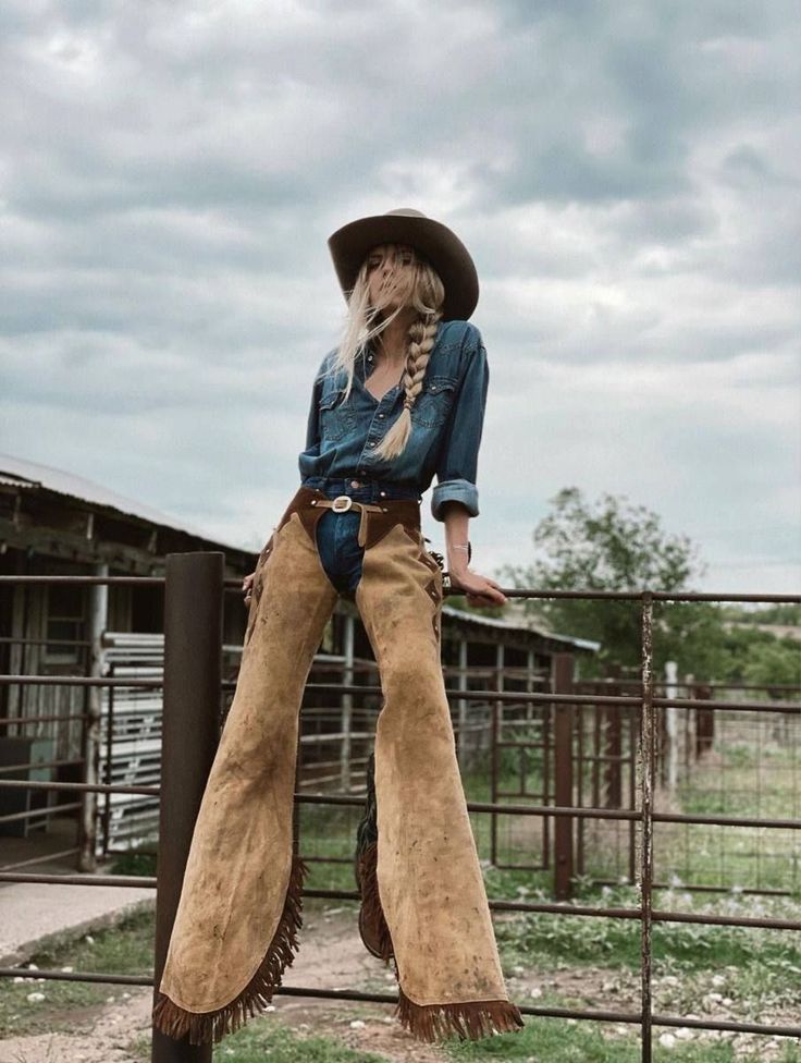 Woman in a cowboy hat and chaps leaning on a fence at a farm