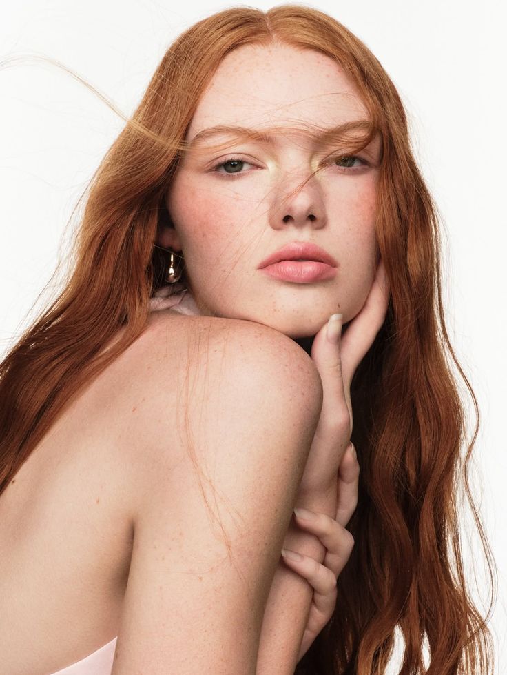 Woman with long red hair and freckles, with exposed shoulders and a hand on her face