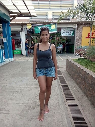 Woman in a tank top and denim shorts standing barefoot on a street