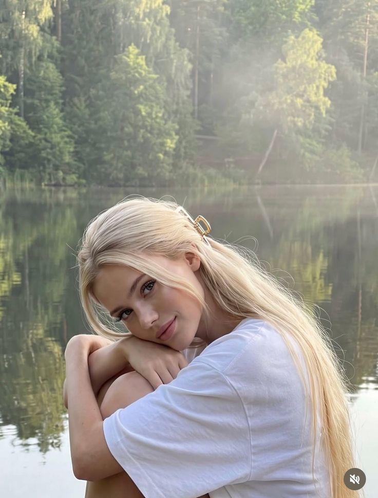 Blonde woman in a white t-shirt posing by a lake with a forest in the background