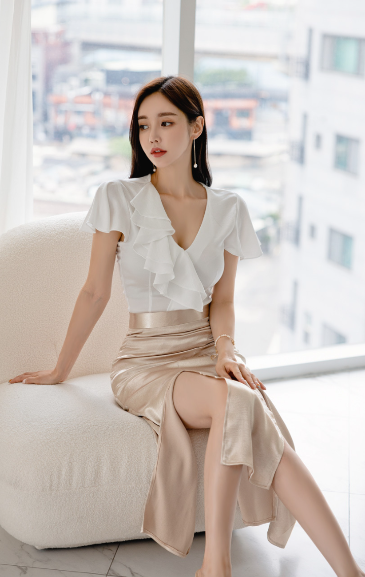 Woman in a white ruffle blouse and a gold slit skirt sitting indoors