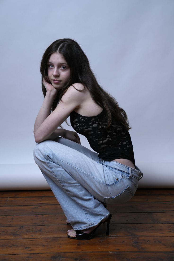 Young woman with long dark hair in a black lace top and jeans, squatting on a wooden floor