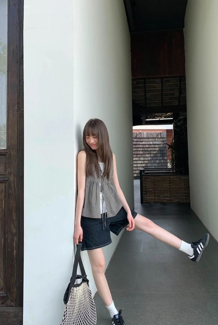 Woman with long hair in denim shorts and a sleeveless top leaning against a wall