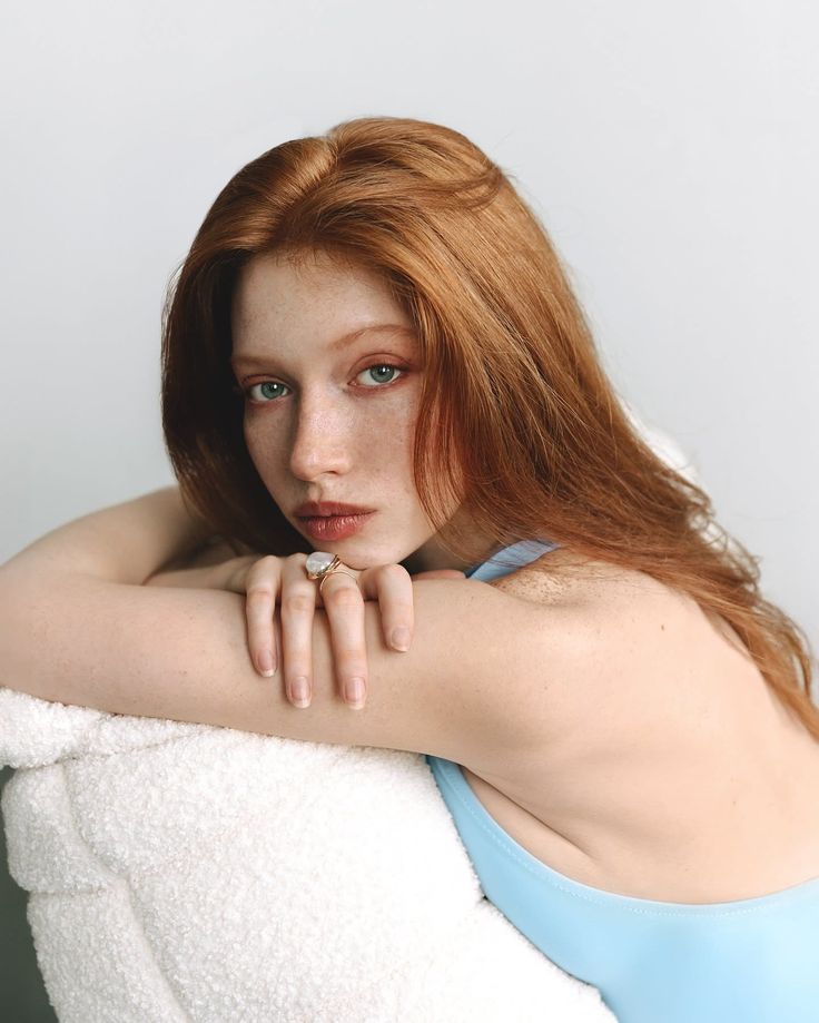Woman with red hair and freckles wearing a light blue top, leaning on a white surface