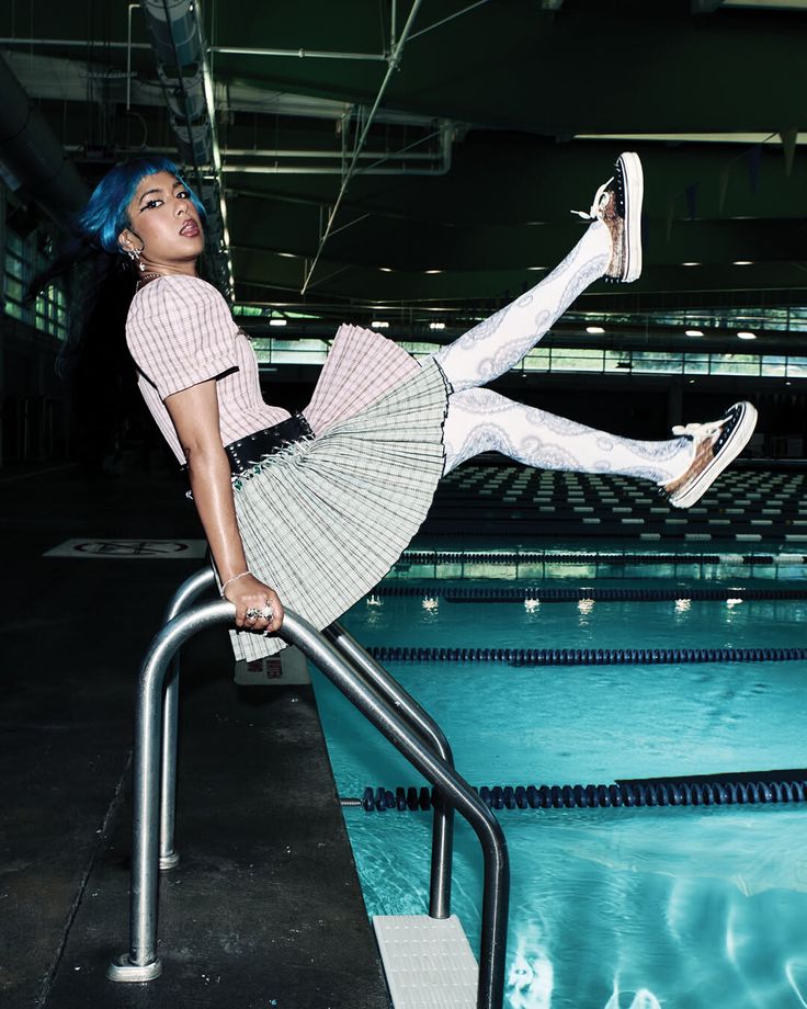Woman with blue hair in a pleated skirt and patterned tights by a swimming pool