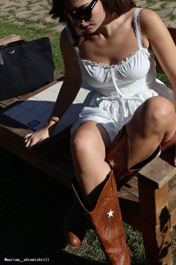 Woman in a white dress and cowboy boots sitting outdoors in bright sunlight
