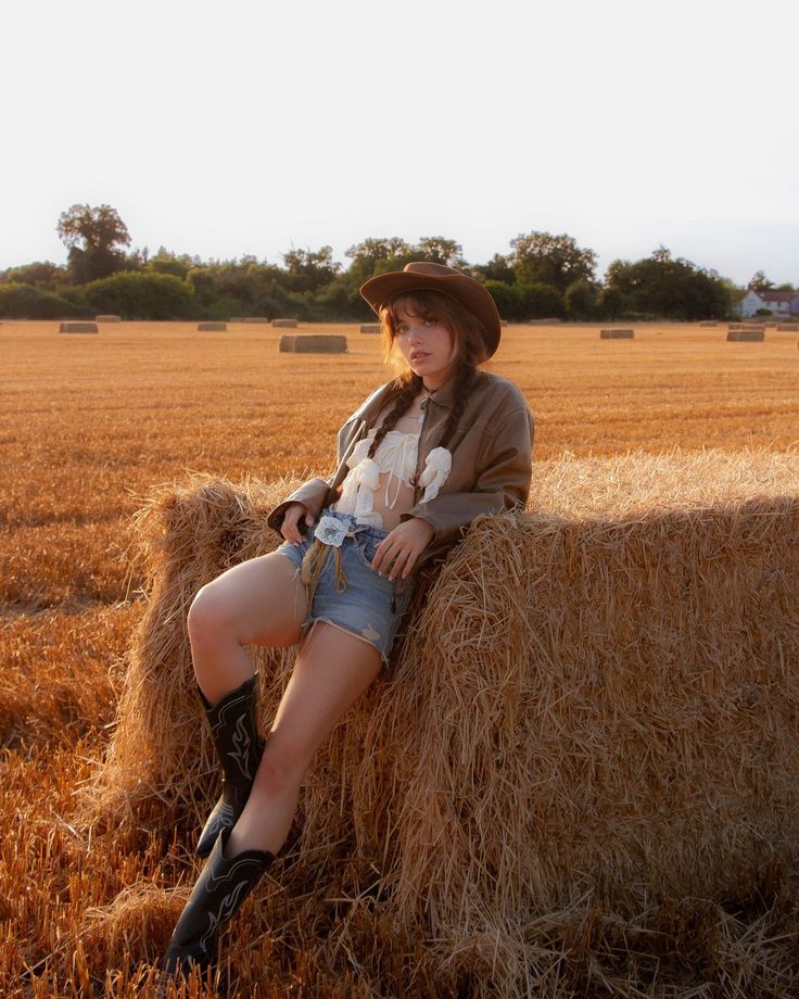 Woman in a cowboy hat and denim shorts leaning on a hay bale at golden hour