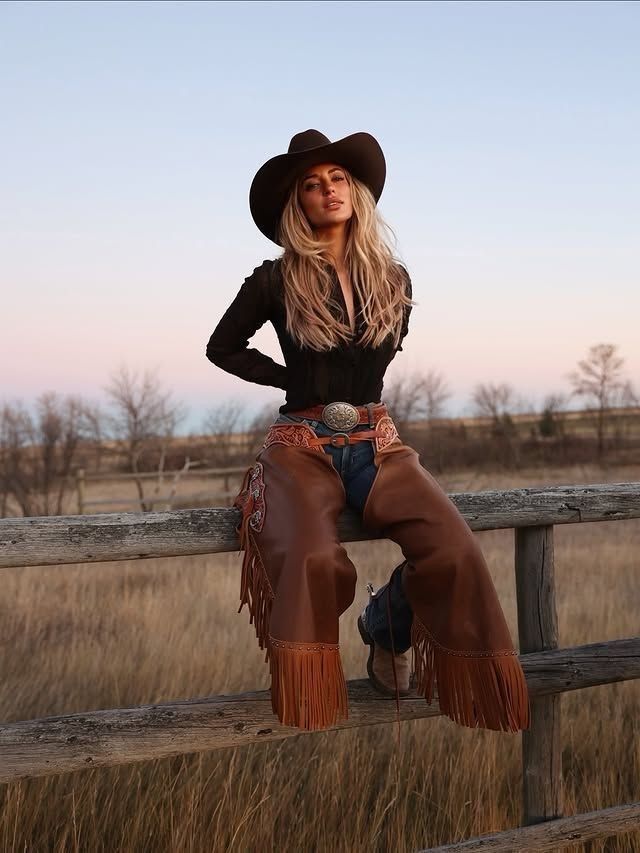 Woman in a cowboy hat and leather chaps sitting on a wooden fence at golden hour