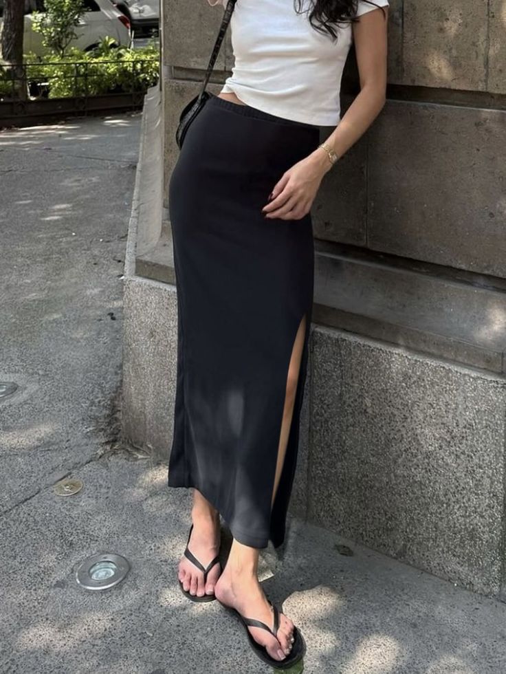 Woman in a black maxi skirt and white crop top leaning against a wall outdoors