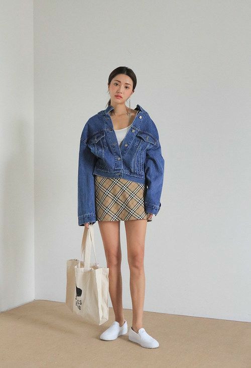 Woman in a denim jacket and plaid skirt holding a tote bag indoors