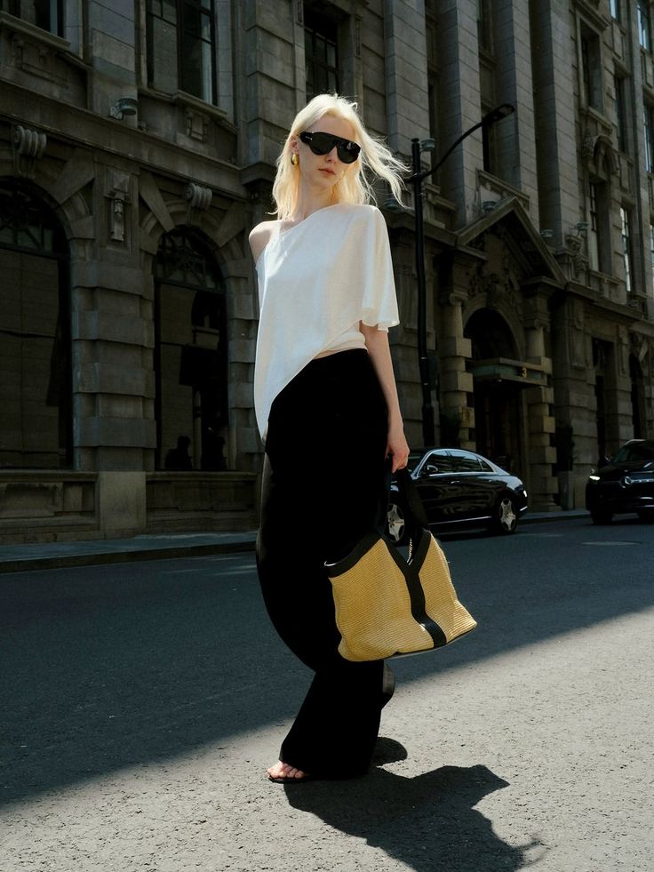 Blonde woman in sunglasses and a white top holding a straw bag on a city street