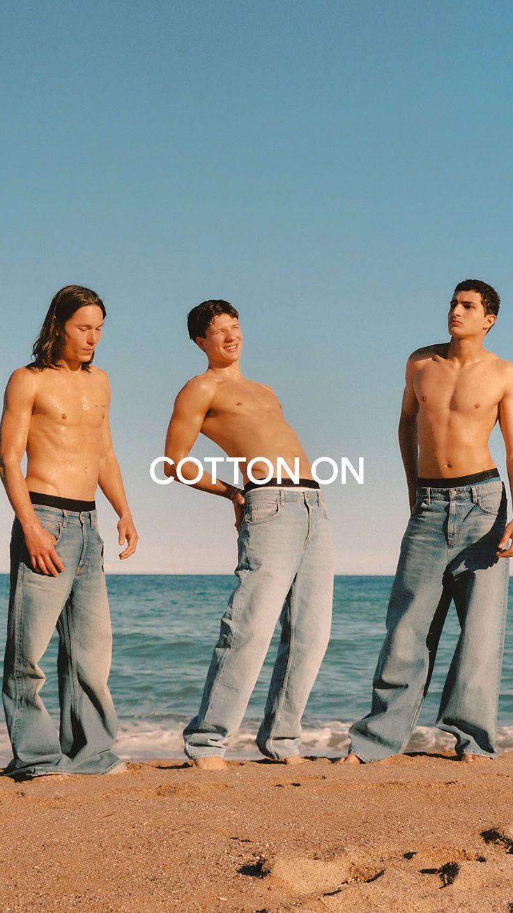 Three shirtless men in jeans on a beach with the ocean in the background