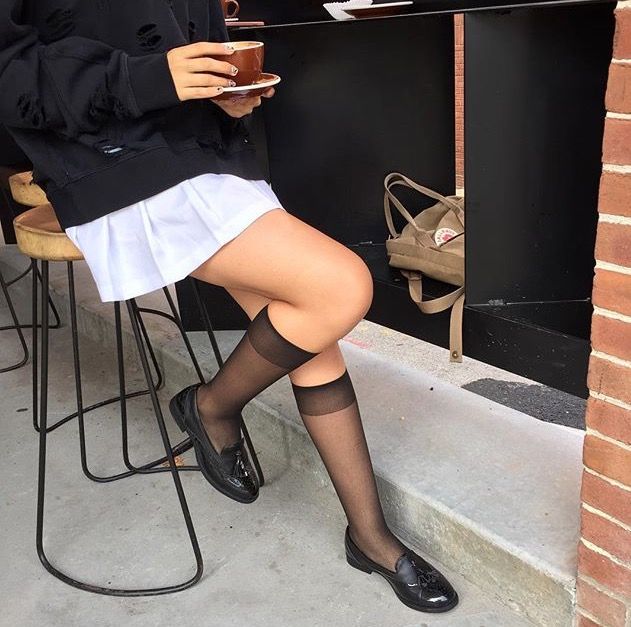 Person in a black hoodie and white skirt sitting outdoors with a coffee cup