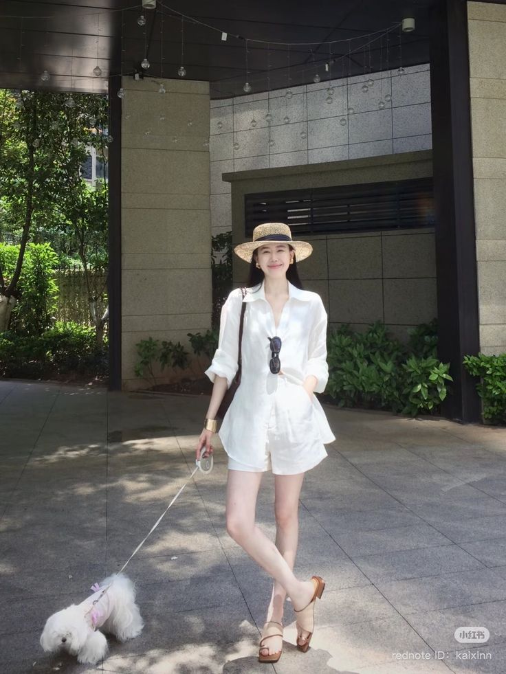Woman in a white shirt and shorts with a straw hat walking a small dog outdoors