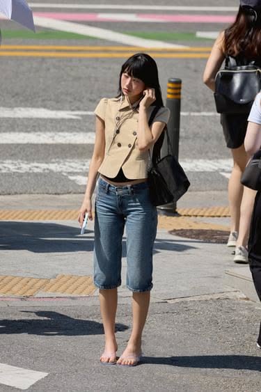 Woman with short black hair wearing a cropped jacket and denim capris, standing barefoot on a street