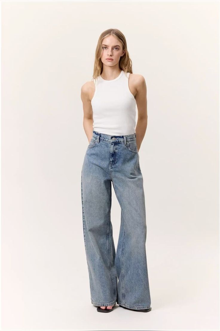 Woman in a white tank top and wide leg jeans posing in a studio