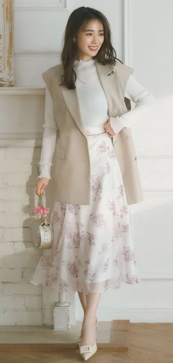 Woman in a floral skirt and sleeveless blazer holding a pink flower indoors