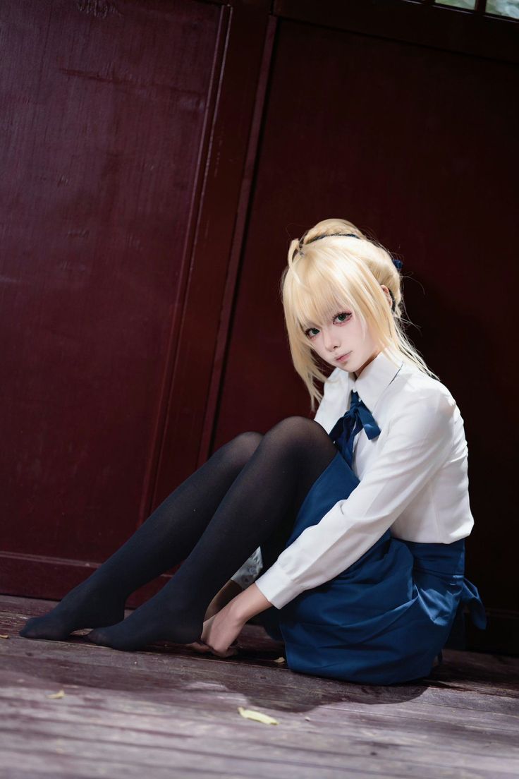 Person with blonde hair in a school uniform and black tights sitting on a wooden floor
