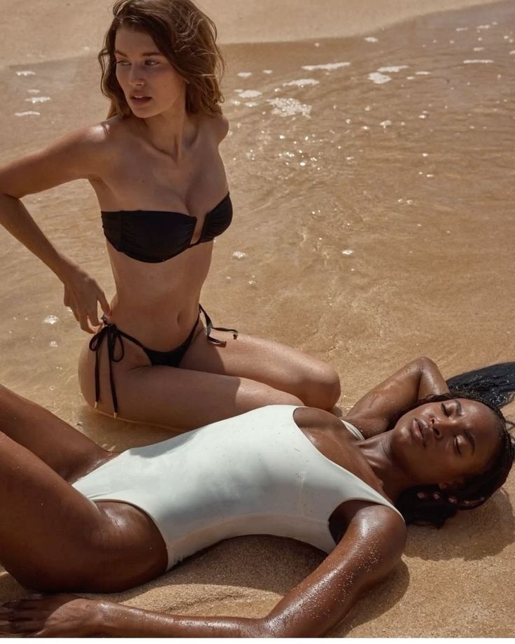 Two women on a sandy beach, one in a bikini and one in a white swimsuit