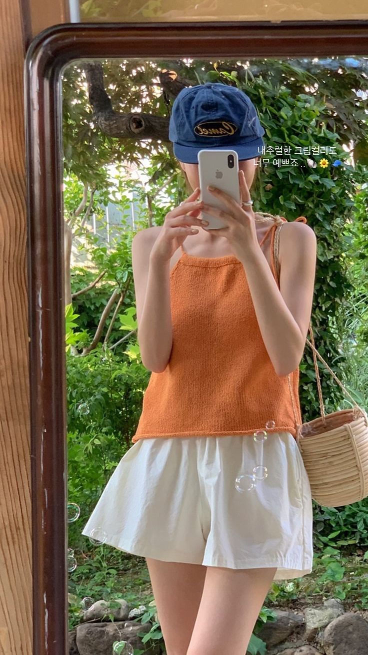 Person in an orange top and white shorts taking a mirror selfie outdoors