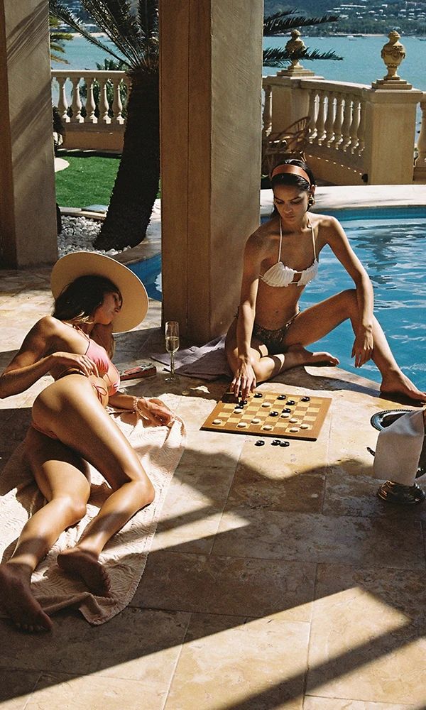 Two women in bikinis playing checkers by a swimming pool on a sunny day