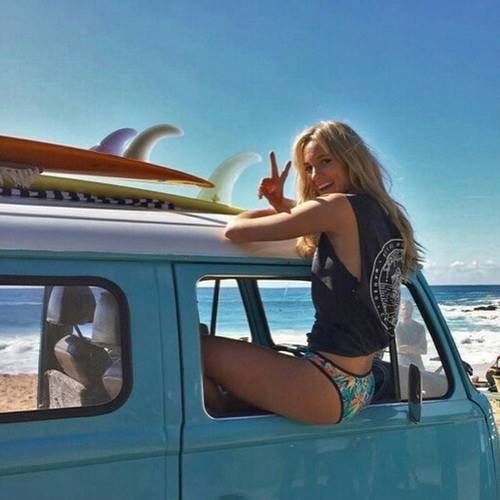 Woman in a bikini and tank top sitting in a van with surfboards on the roof at the beach