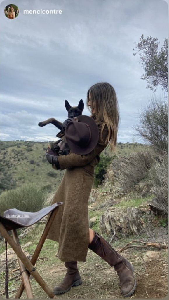 Woman in a brown dress and boots holding a dog and a hat in a hilly outdoor setting
