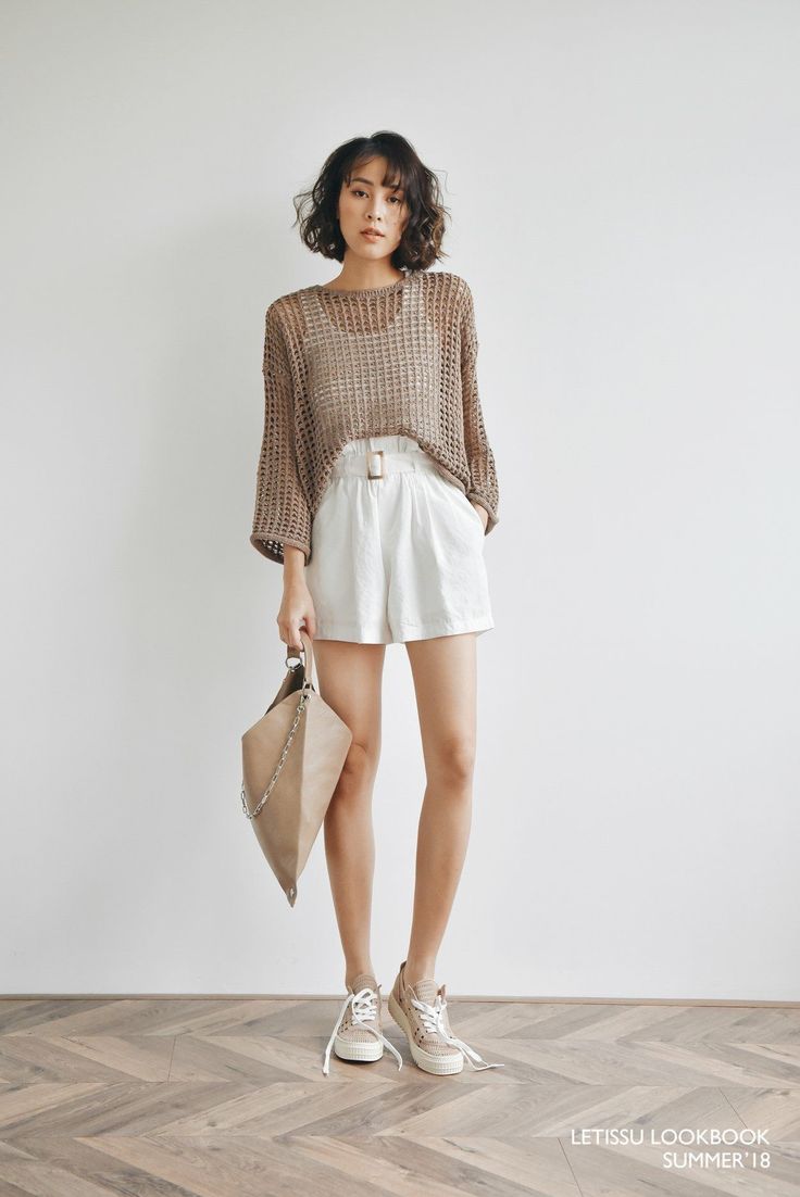 Woman with short hair wearing a brown knit top and white shorts, holding a handbag