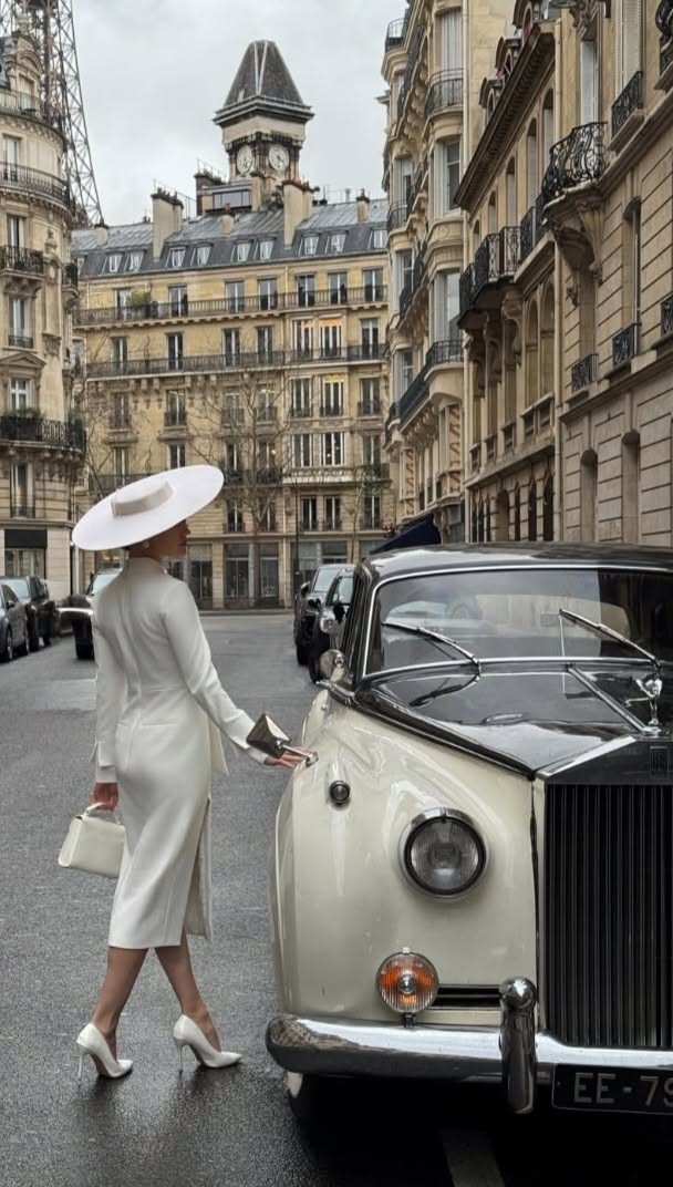 Woman in a white dress and hat next to a vintage car on a city street