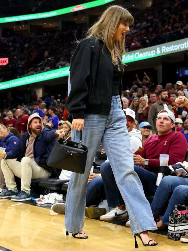 Woman in a black bomber jacket and blue jeans walking past a crowd indoors