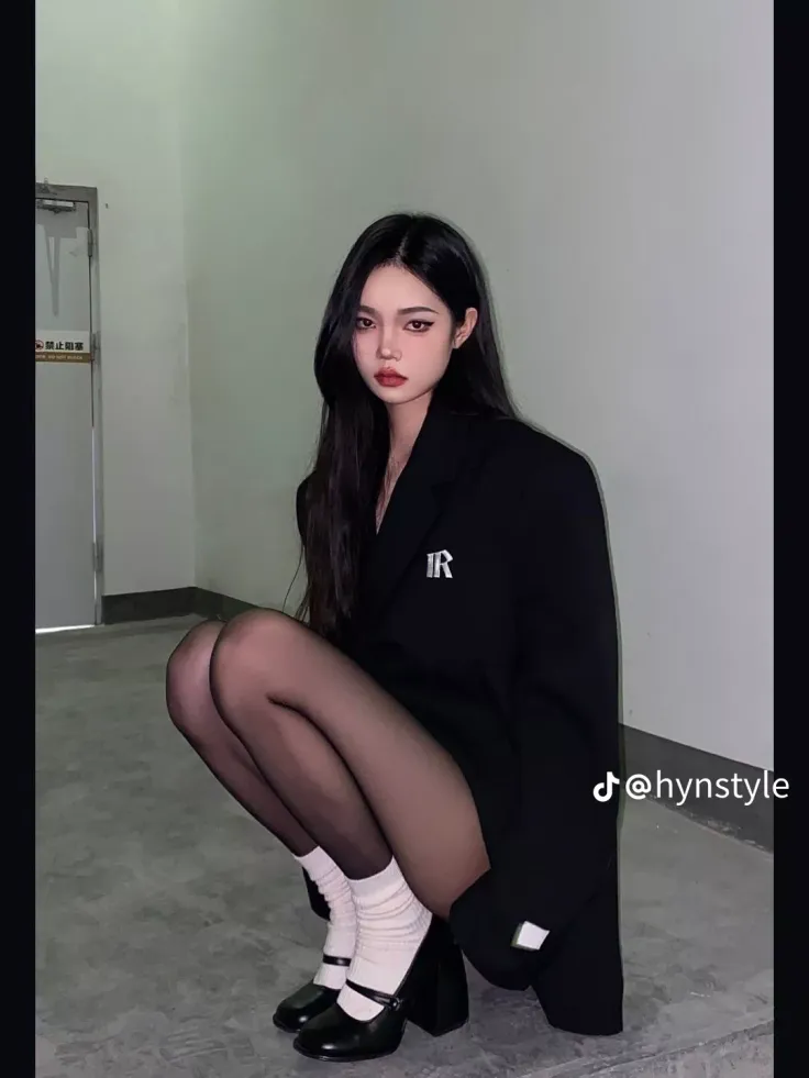 Woman with long black hair wearing a black blazer and tights, squatting indoors