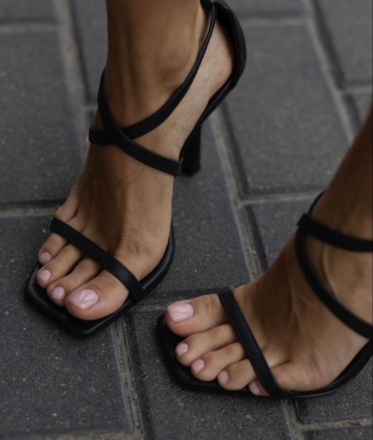 Close up of feet in black high-heeled sandals with pink nail polish on paved ground