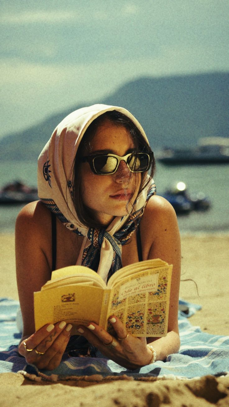 Woman in a head scarf and sunglasses reading a book on the beach