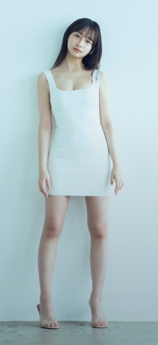Woman in a white dress with long dark hair standing against a light wall