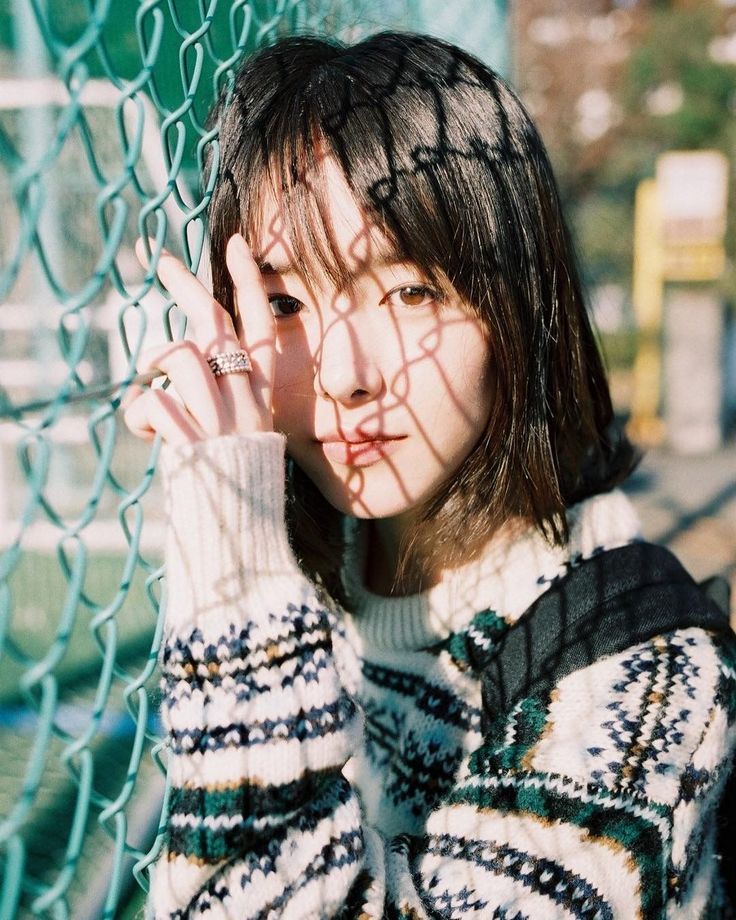 Woman with short hair and a fair isle sweater behind a chain link fence with shadows on her face