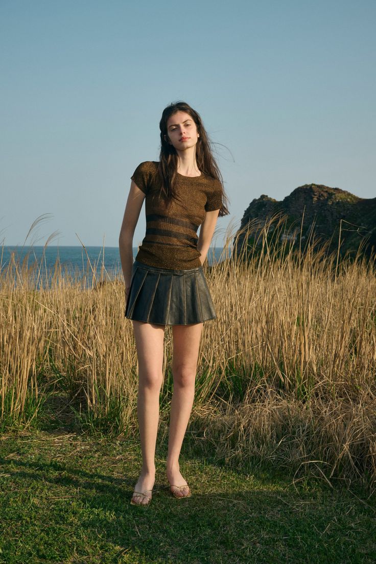 Woman in a dark top and mini skirt standing in tall grass by the ocean