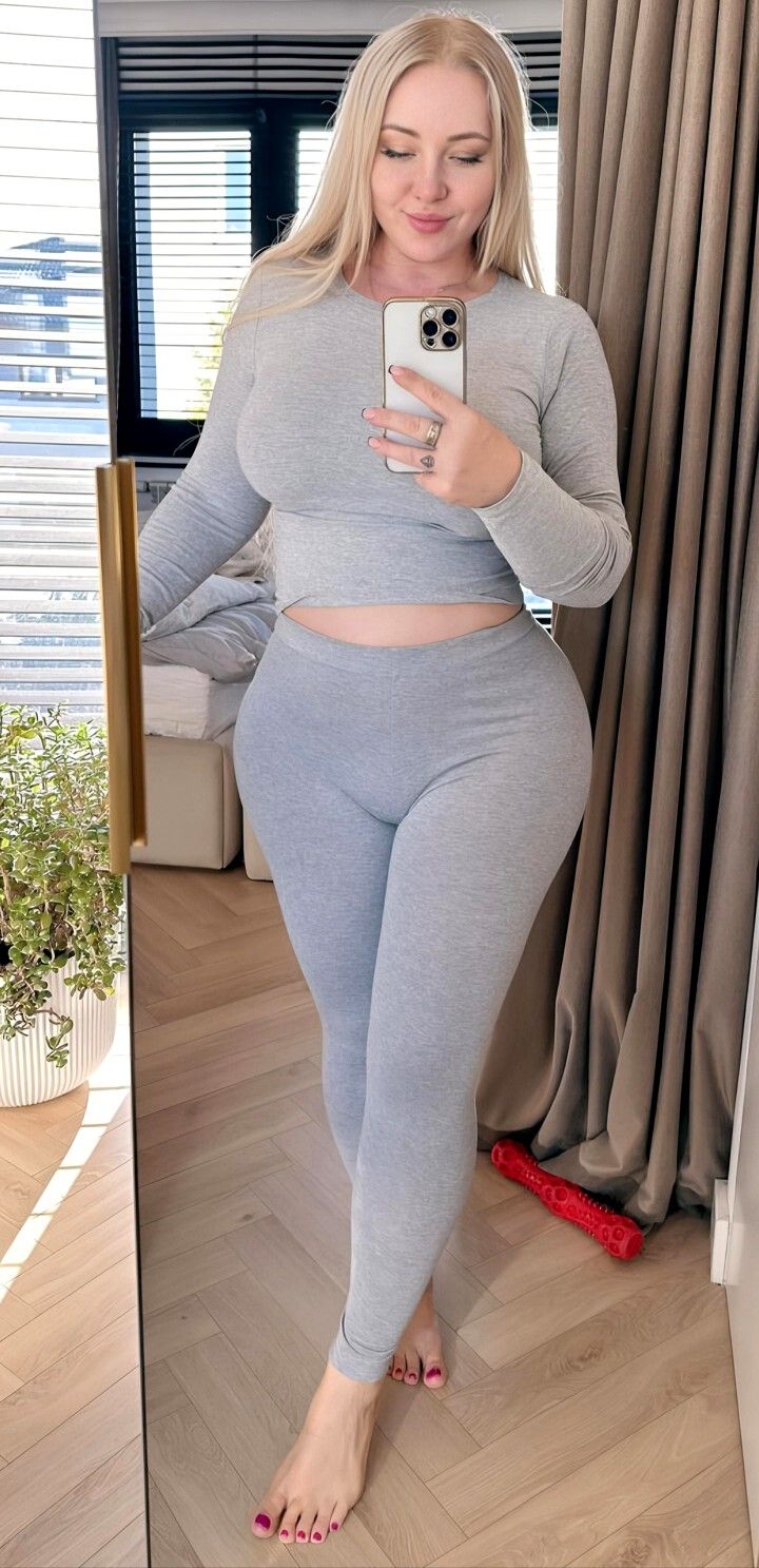 Woman with long blonde hair in a grey crop top and leggings taking a mirror selfie