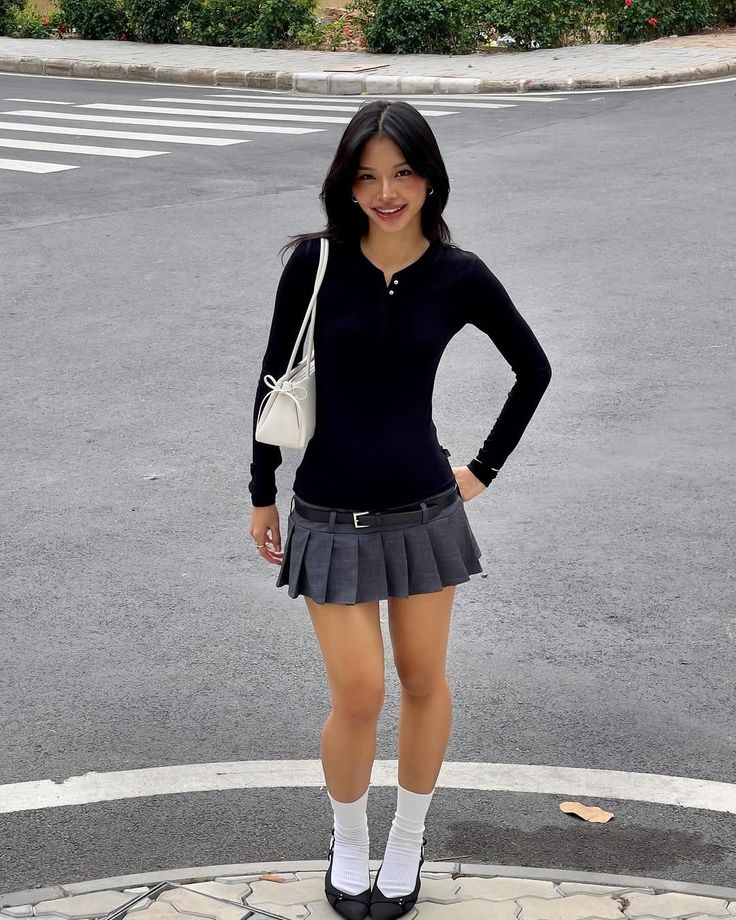 Woman in a black long-sleeve shirt and gray pleated skirt standing outdoors