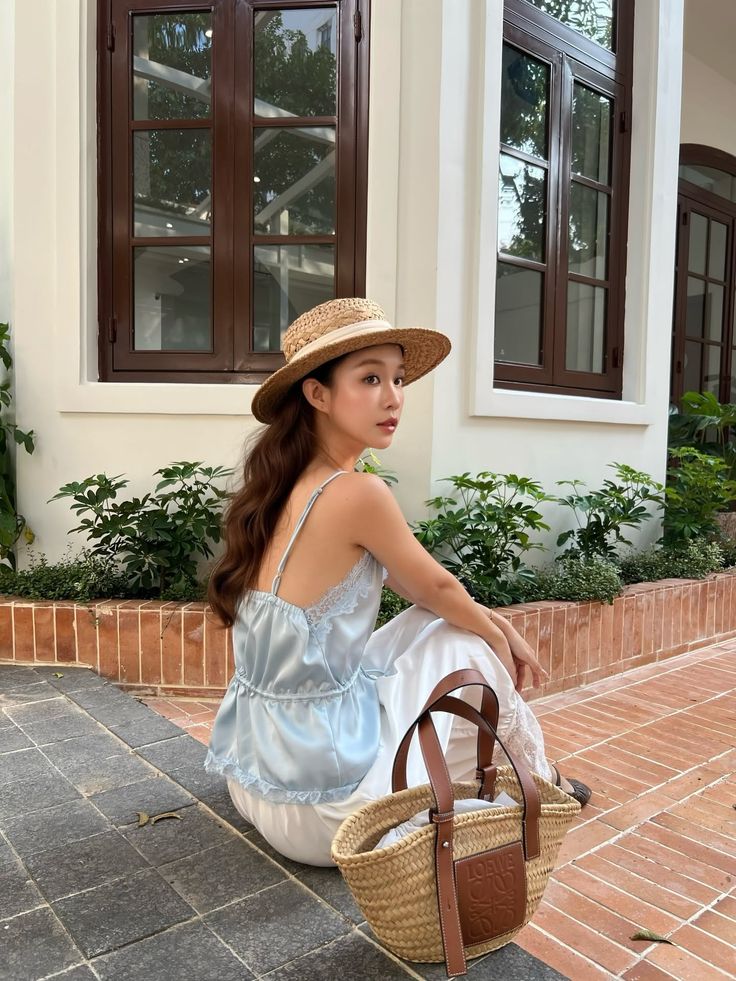 Woman in a straw hat and light blue top sitting outdoors with a straw bag