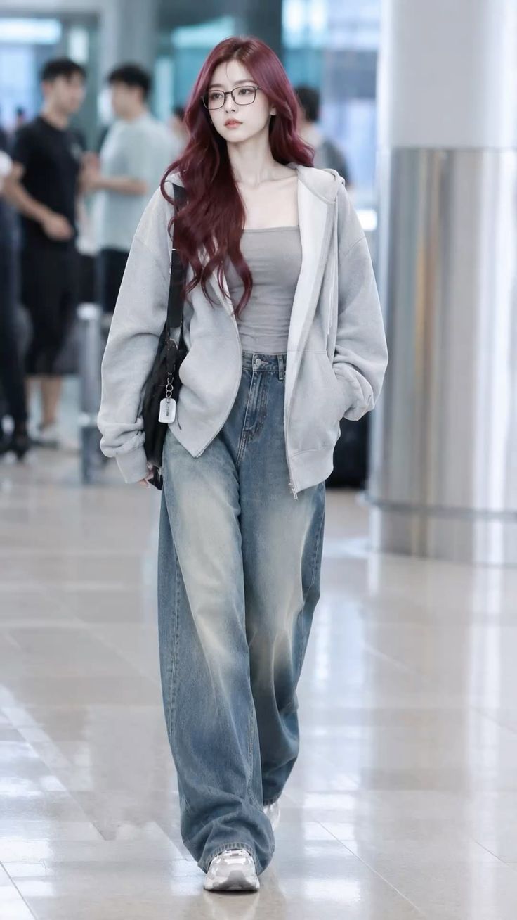 Woman with long red hair and glasses wearing a gray hoodie and baggy jeans indoors