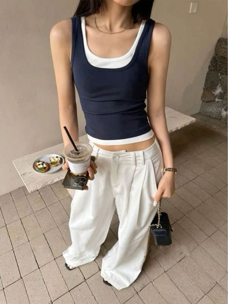 Person in a layered tank top and white pants holding a coffee cup indoors