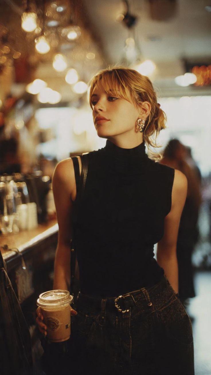 Woman with blonde hair and a black turtleneck top holding a drink indoors