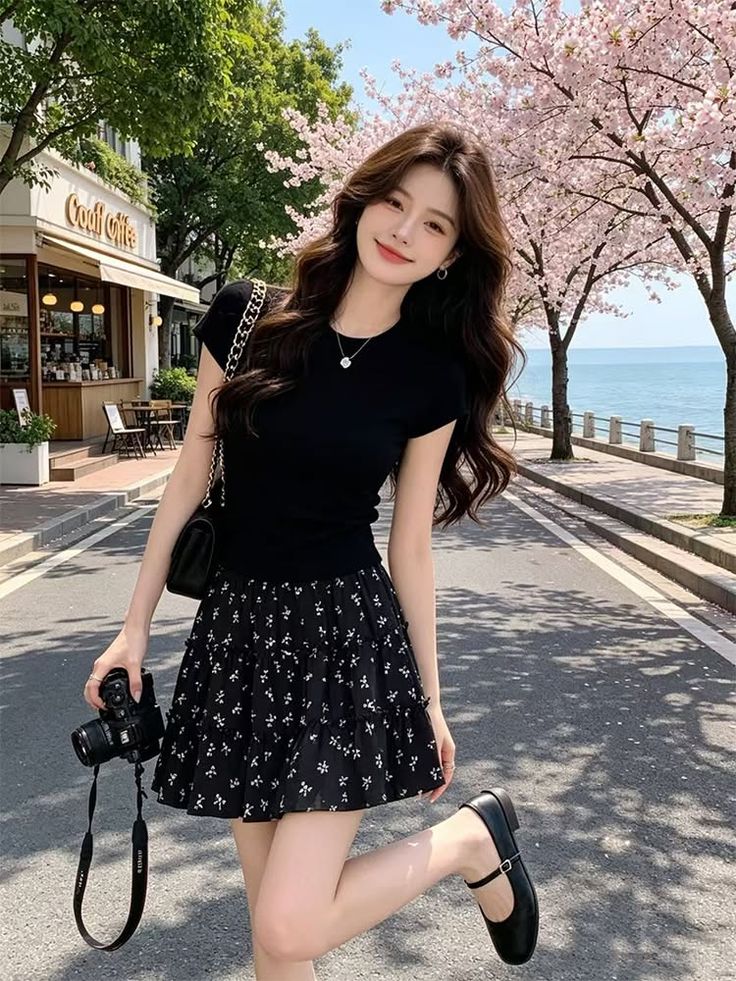 Woman in a black top and floral mini skirt holding a camera on a street with cherry blossoms