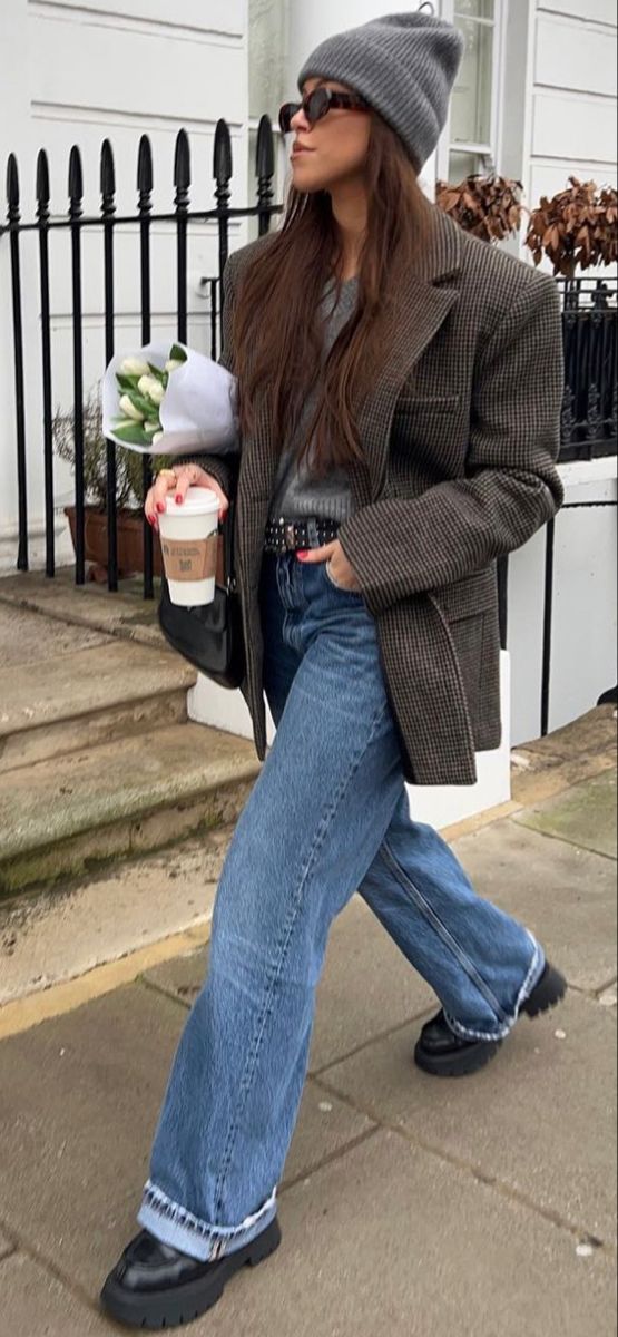Woman in a gray beanie and plaid blazer walking outdoors with coffee and flowers